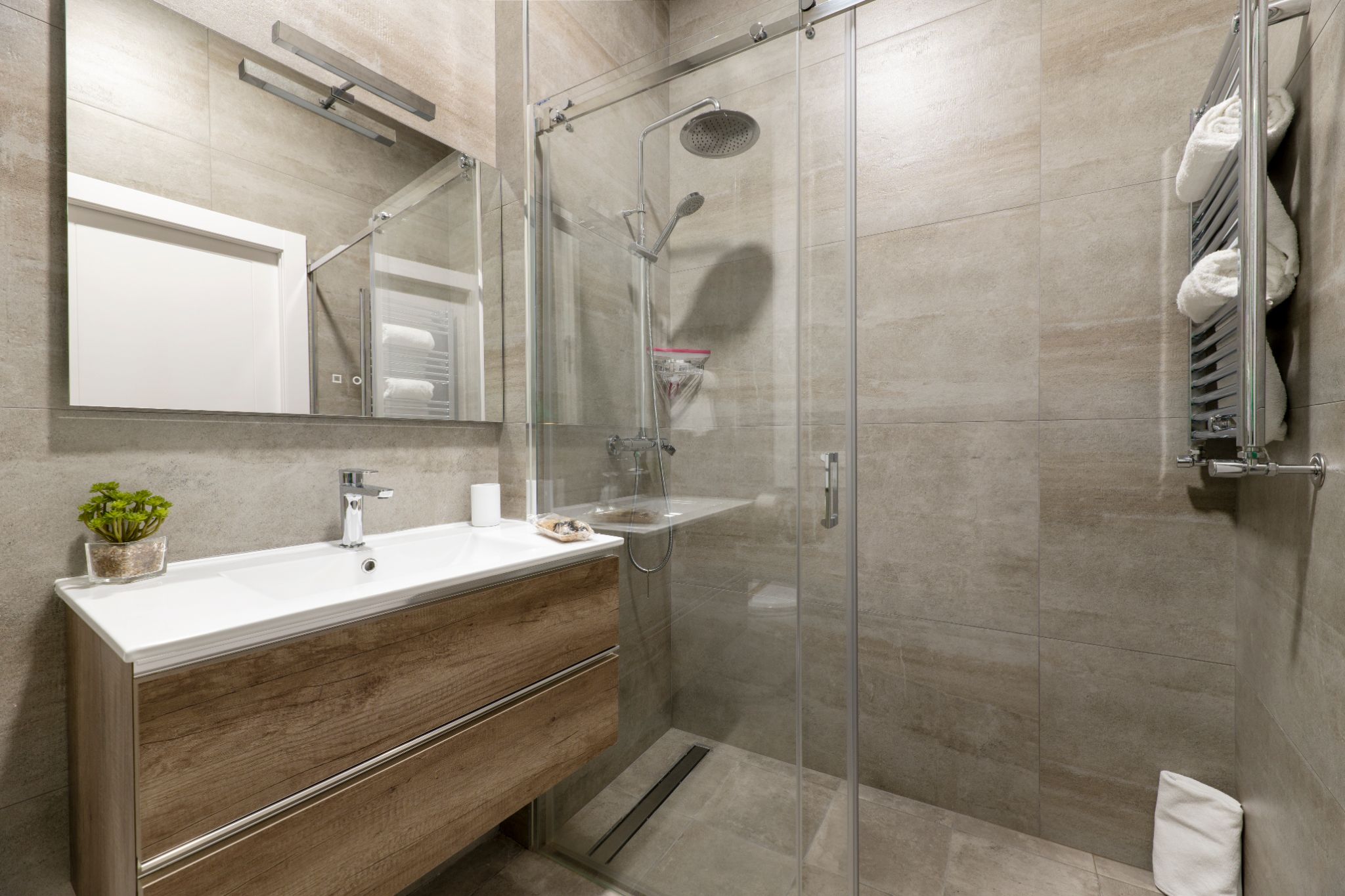 Modern bathroom with wooden floating vanity, large mirror, glass sliding shower door, and neutral stone tiling.