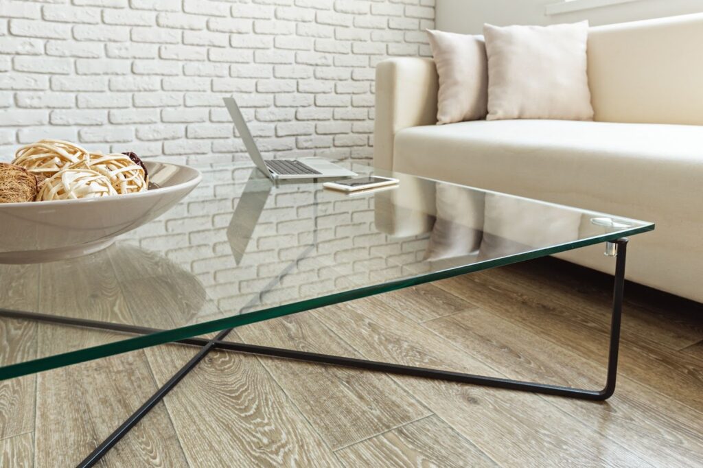 A modern living room with a glass coffee table, laptop, smartphone, decorative bowl, and a beige sofa against a white brick wall.