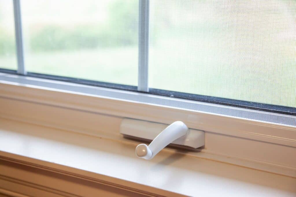 Close-up of a white window crank handle with a screen and greenery outside.