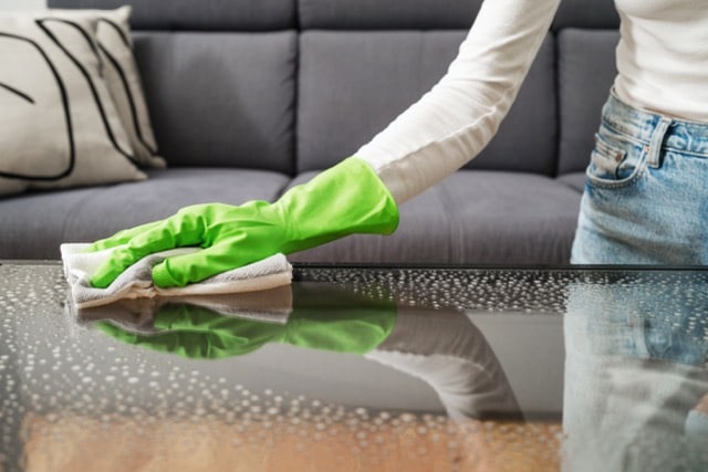 Woman wearing green gloves cleans a glass table with a cloth, focusing on removing smudges and dirt.