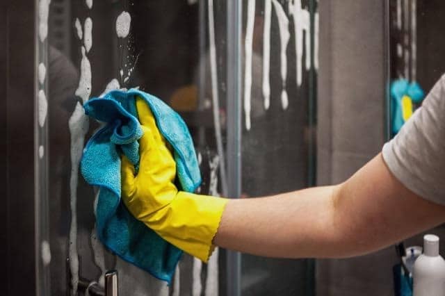 A person in yellow gloves wipes a glass shower door ensuring it sparkles in a bright bathroom environment