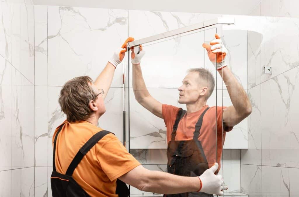 Two men in orange overalls are repairing a shower door in a bathroom setting