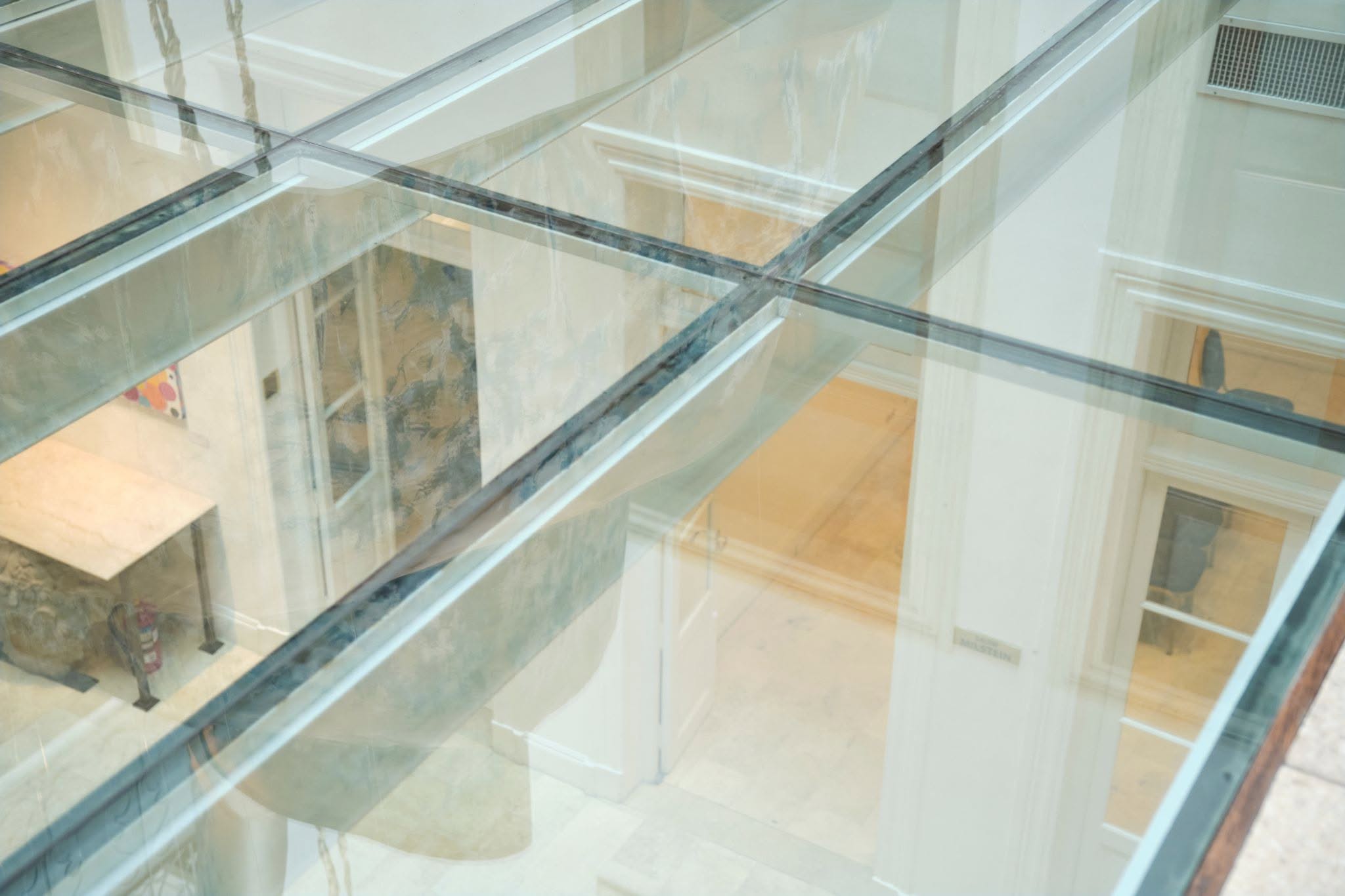 High-angle view of a structural glass floor walkway within a modern multi-level building, revealing the architectural details of the floor below.