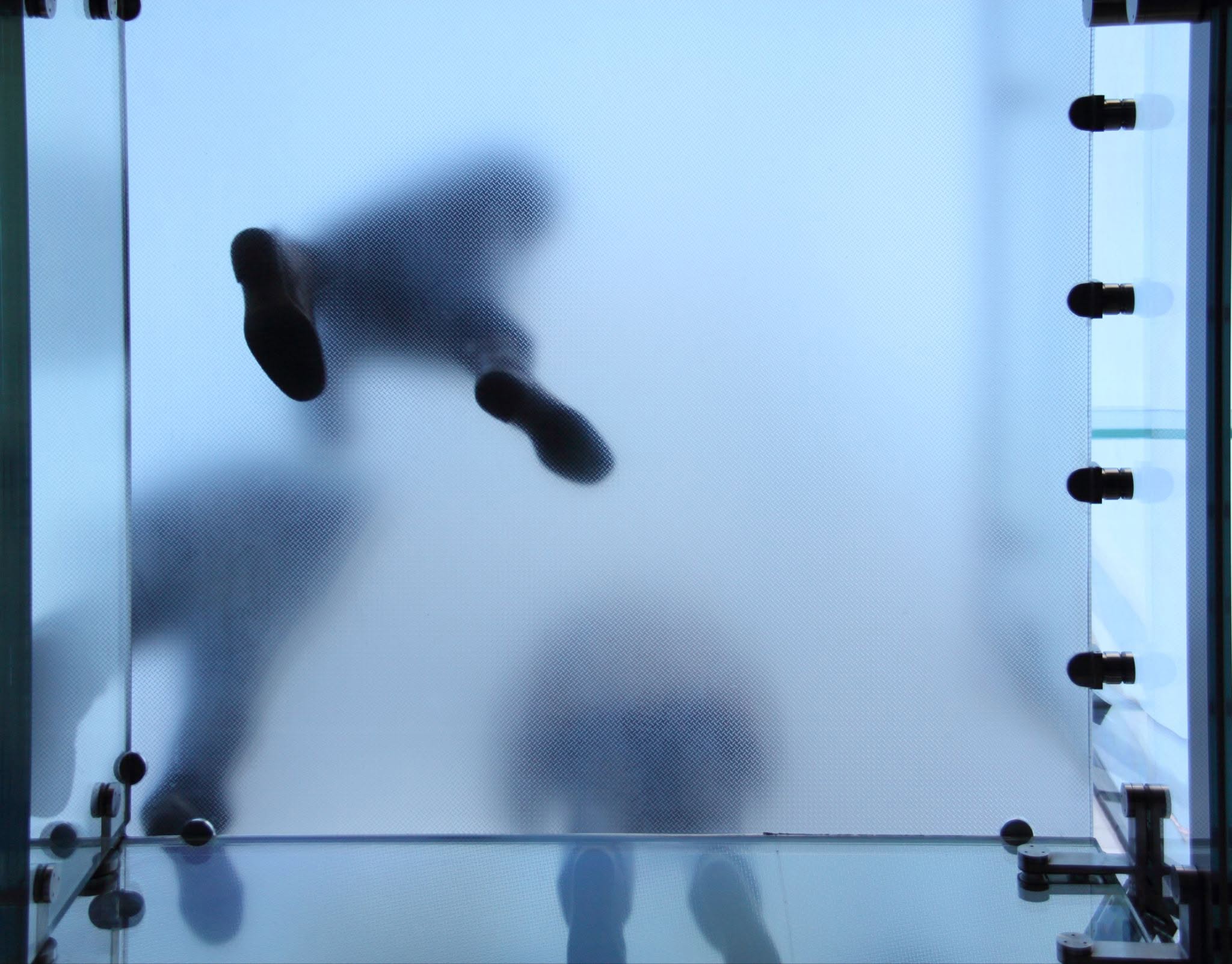 Low-angle silhouette of people walking across a textured, translucent glass floor panels secured with metal hardware.