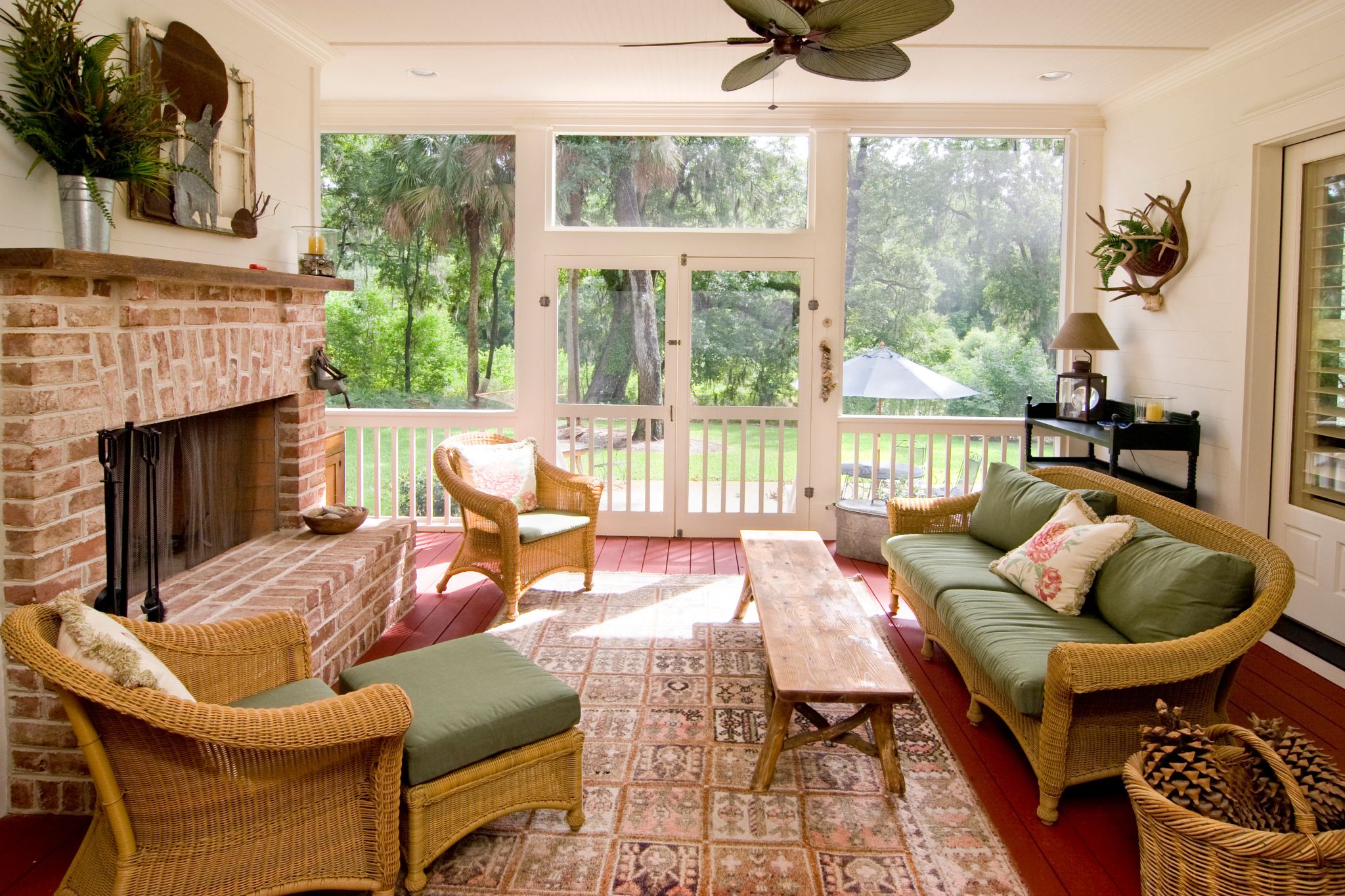 Cozy screened porch featuring wicker furniture, green cushions, a brick fireplace, and large windows overlooking trees.