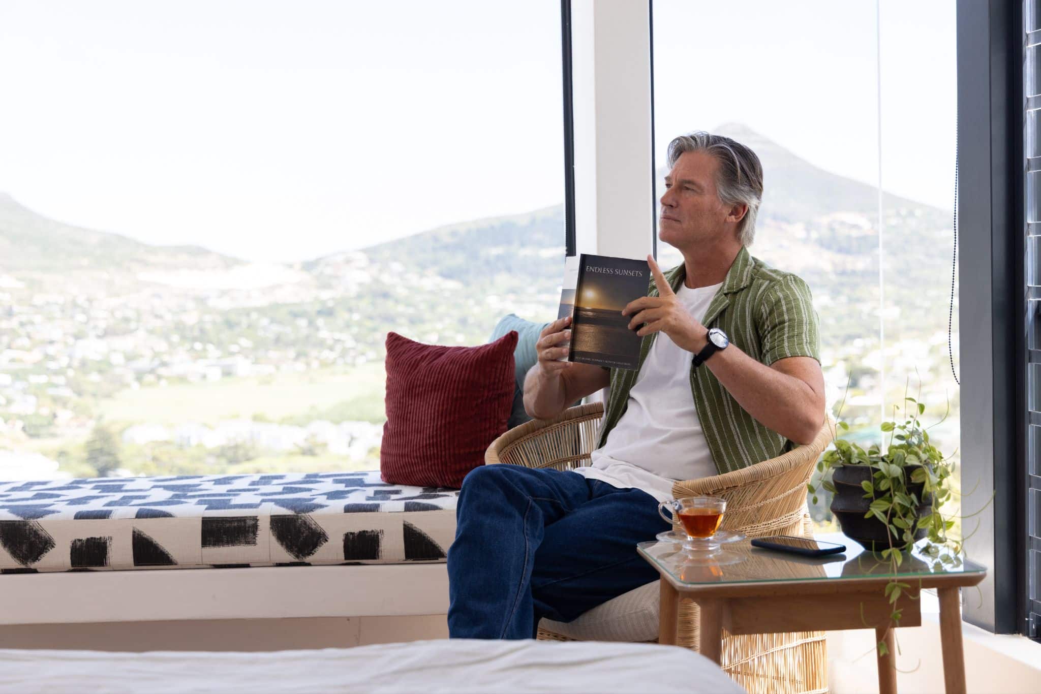 Man holding a book while sitting in a wicker armchair near a large window with scenic mountain views.