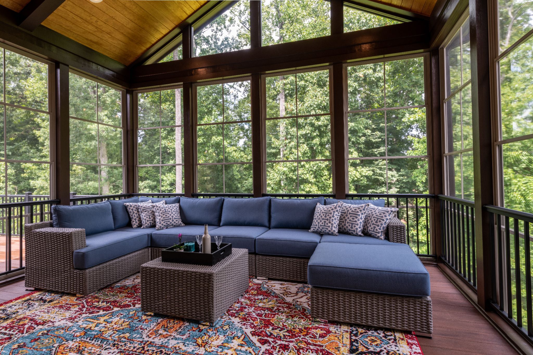 Spacious screened porch with blue outdoor sectional sofa, patterned rug, and wooden ceiling overlooking a forest.