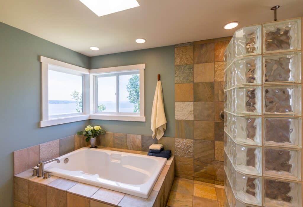 Modern bathroom featuring a soaking bathtub surrounded by tan tile, corner windows, and a glass block shower wall.