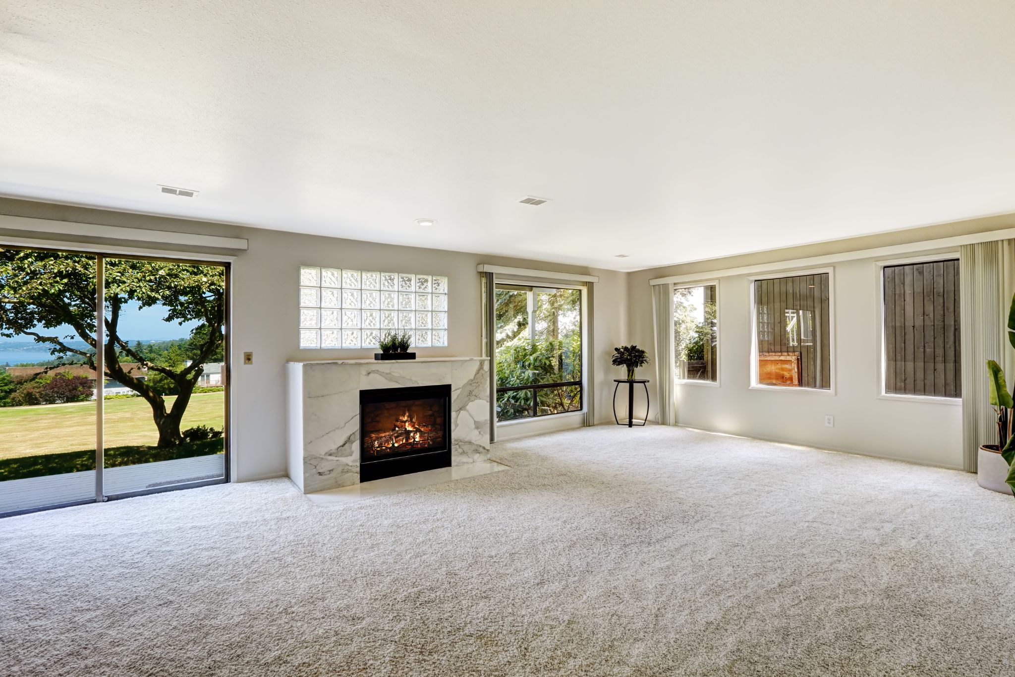 Spacious, empty living room with light carpet, a marble fireplace, a glass block accent window, and large sliding doors leading outside.