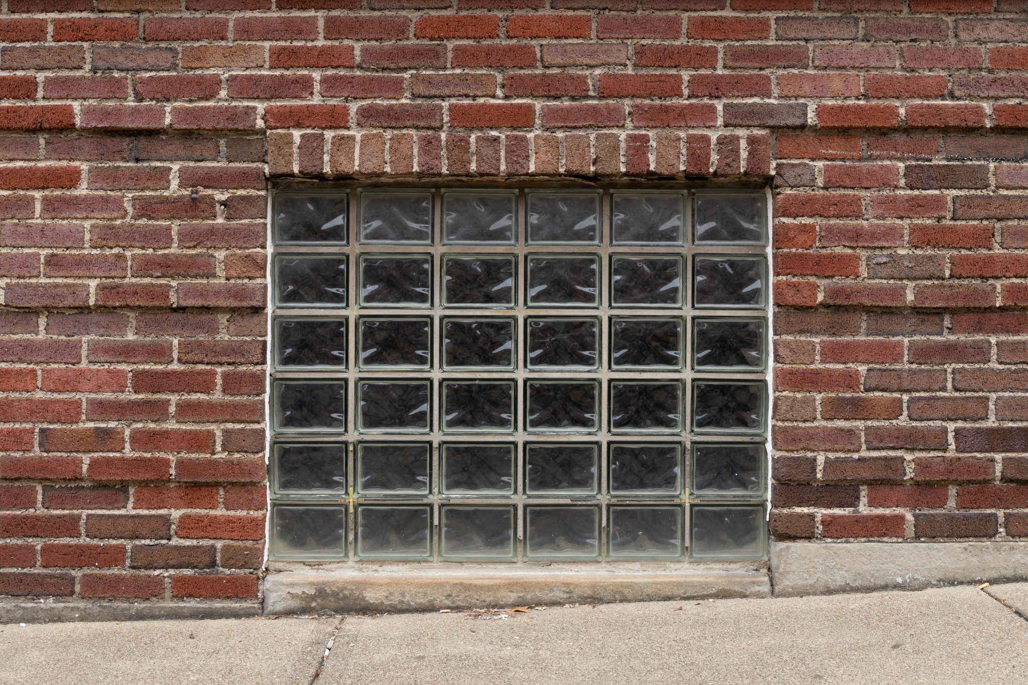 Square glass block window set into a red brick wall above a concrete sidewalk.