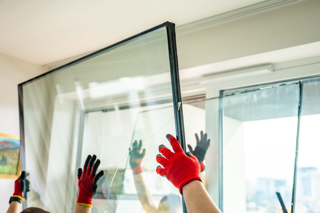 Gloved workers carefully lifting a large pane of window glass for installation indoors.