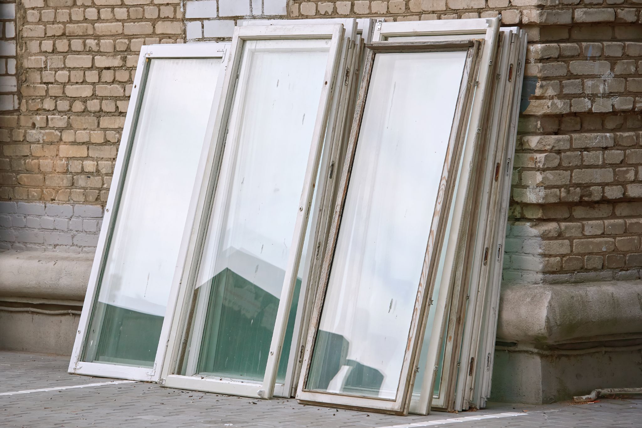 Several old white window frames with glass panes leaning against a brick building wall.