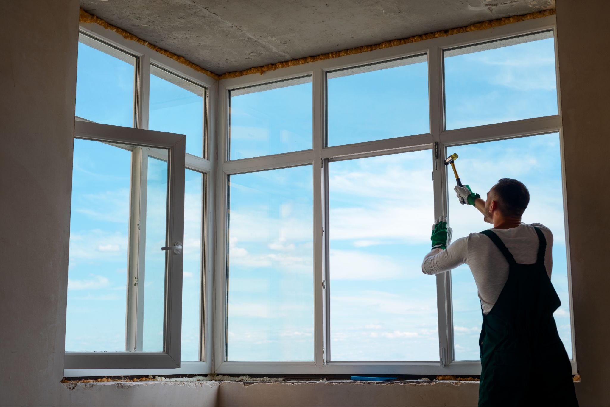 Worker in overalls using a mallet to install new white vinyl windows overlooking a blue sky.